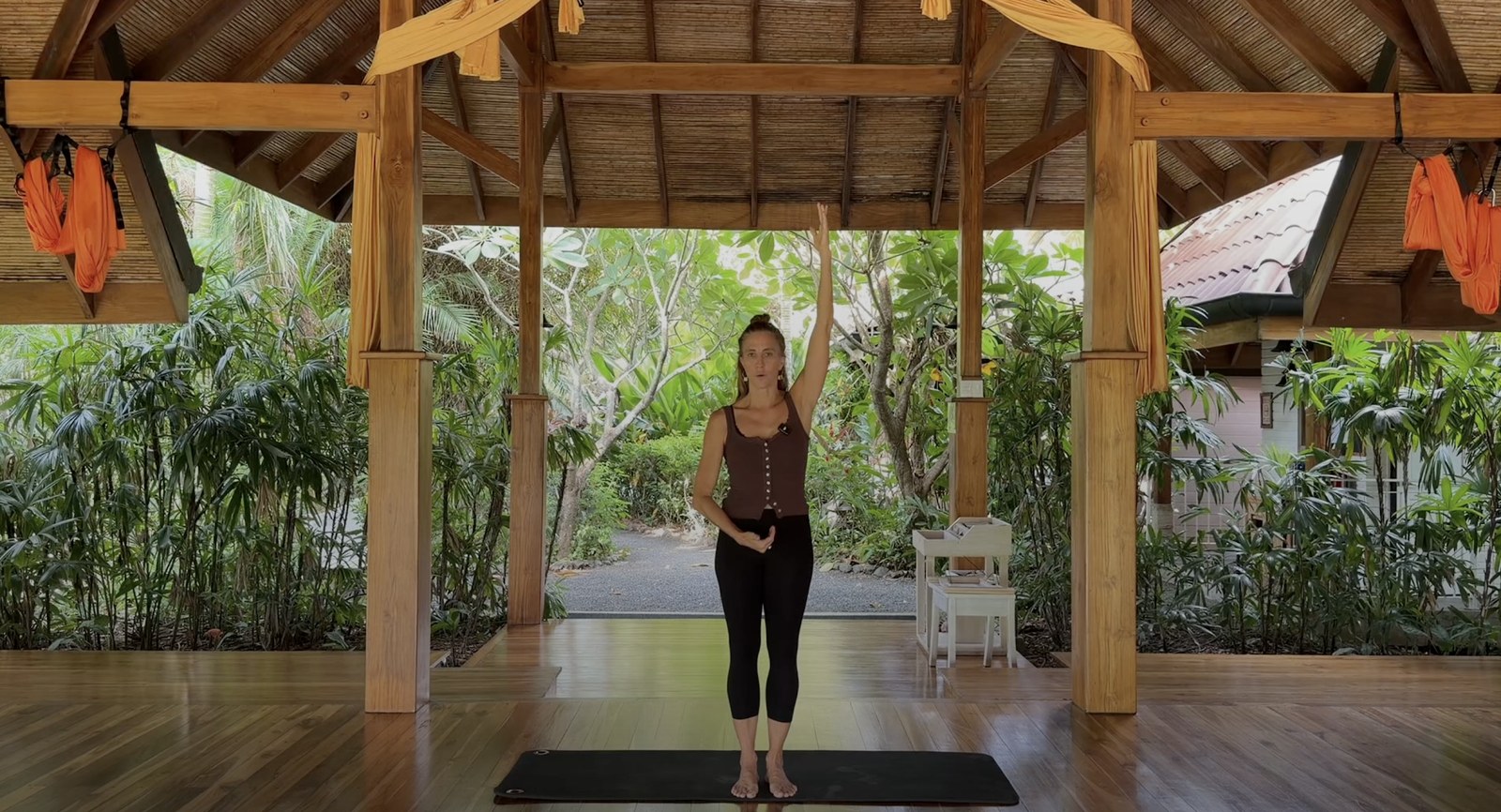 Pilates instructor demonstrating a standing movement with one arm raised in an open-air studio